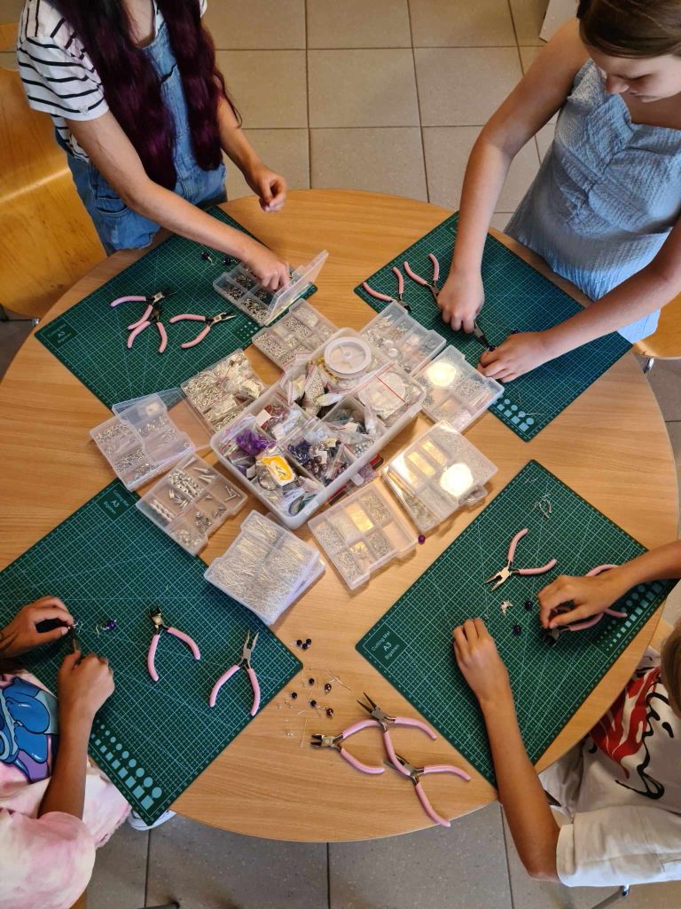 grupa dziewczynek siedzi przy stole na którym leżą maty samogojące oraz narzędzia i pudełka z półproduktami do tworzenia biżuterii