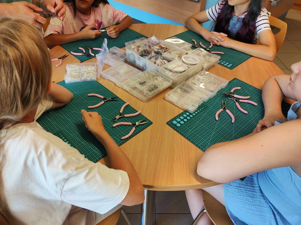 grupa dziewczynek siedzi przy stole na którym leżą maty samogojące oraz narzędzia i pudełka z półproduktami do tworzenia biżuterii
