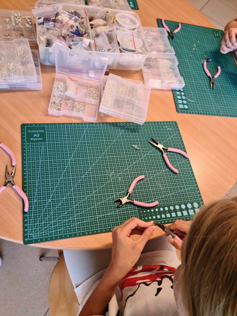 dziewczynka siedzi przy stole na którym leżą maty samogojące oraz narzędzia i pudełka z półproduktami do tworzenia biżuterii