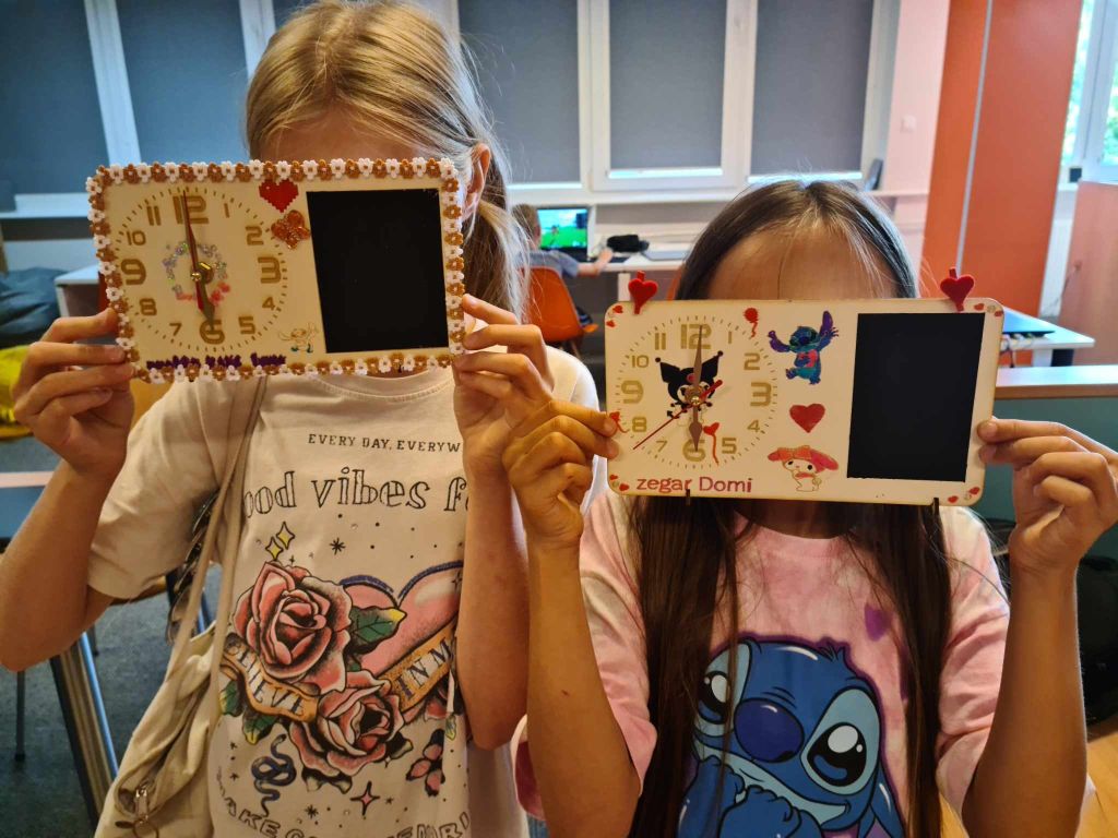 Dwie dziewczynki pokazują do zdjęcia własnoręcznie wykonane tabliczki z zegarami