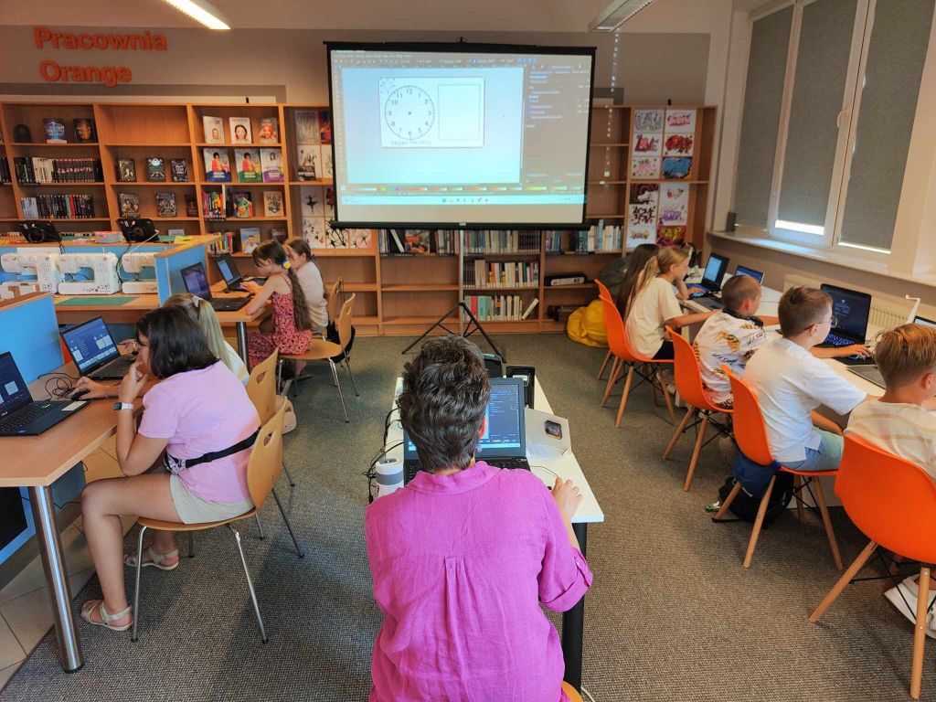 grupa dzieci siedzi przy biurkach przed laptopami, po środku kobieta przy laptopie, w tle ekran z wyświetlanym obrazem