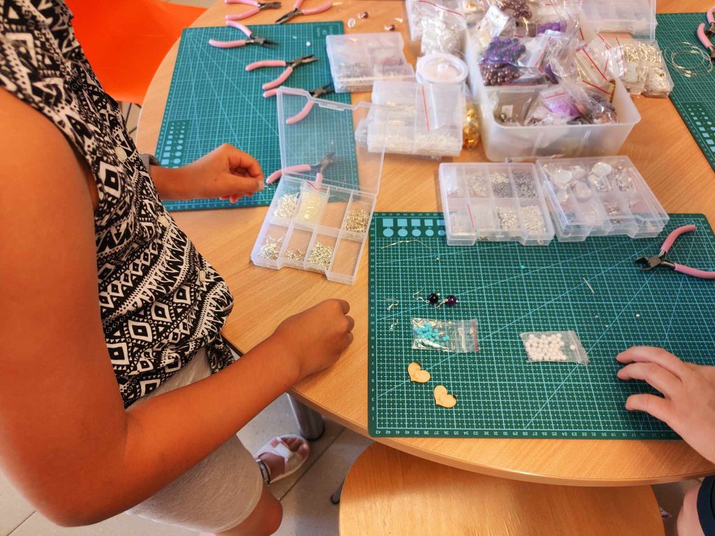 dziewczynka stoi przy stole na którym leżą maty samogojące oraz narzędzia i pudełka z półproduktami do tworzenia biżuterii