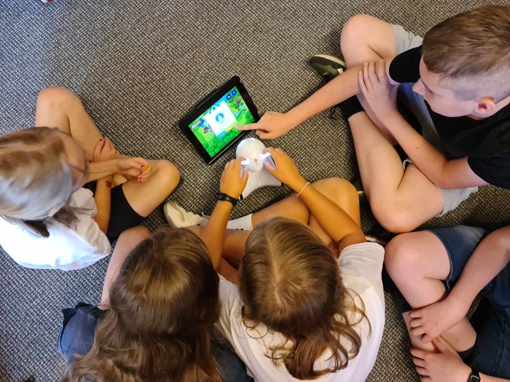 Dzieci siedzą w grupie na wykładzinie, przed nimi stoi tablet i robot Photon
