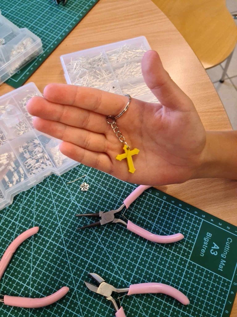 ręka prezentująca mały wisiorek z druku 3D w kształcie krzyża, w tle maty samogojące oraz narzędzia i pudełka z półproduktami do tworzenia biżuterii