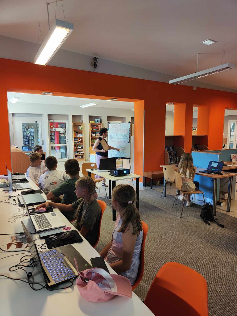 grupa dzieci siedzi przy laptopach, po środku stoi kobieta