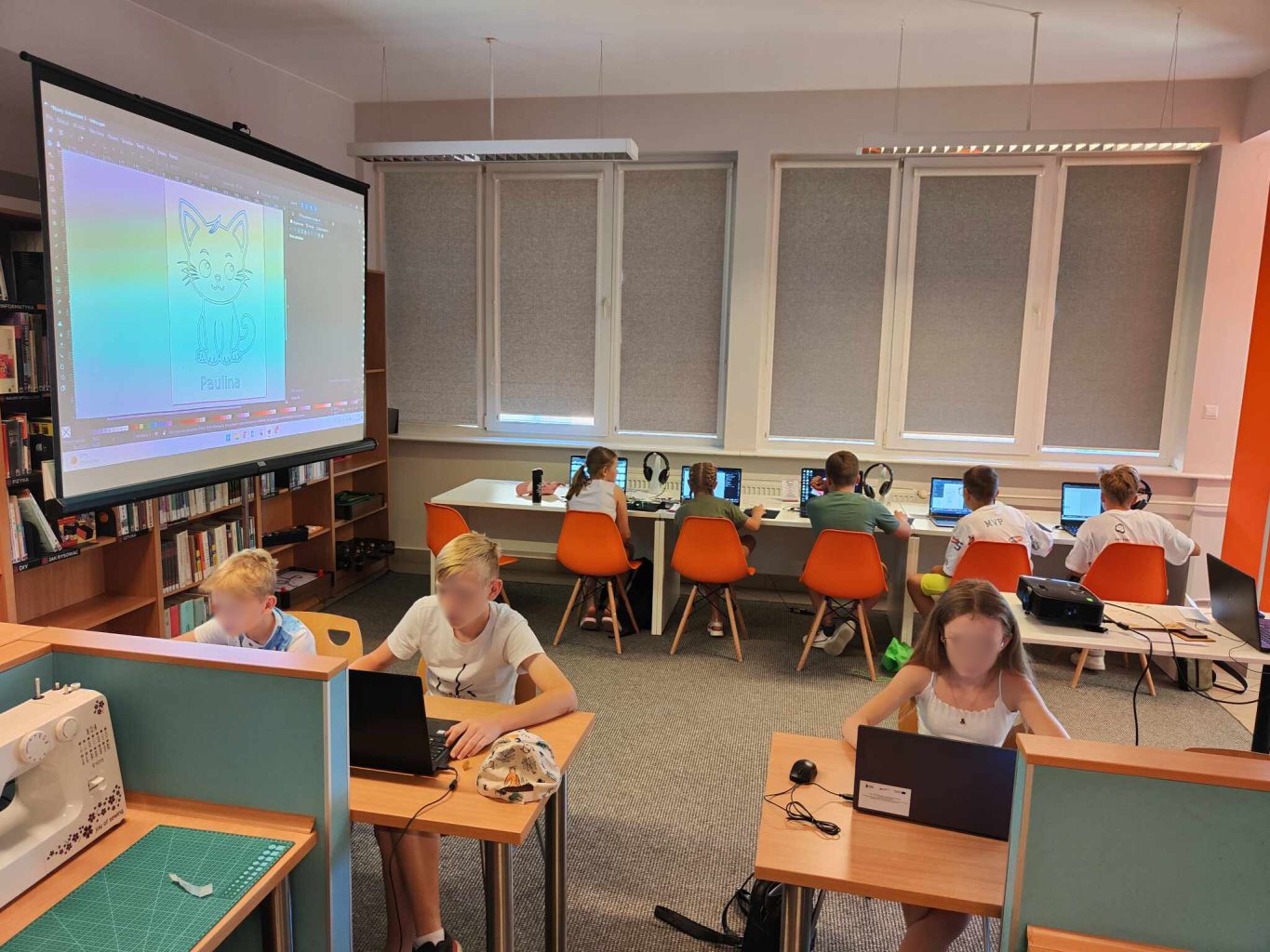 grupa dzieci siedzi przy biurkach przed laptopami, z lewej ekran