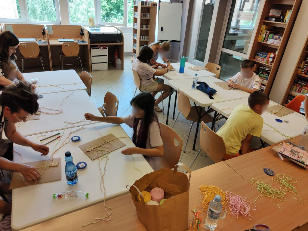 Grupa dzieci siedzi przy stołach ustawionych obok siebie. Po lewej nad jedną z osób pochyla się kobieta.