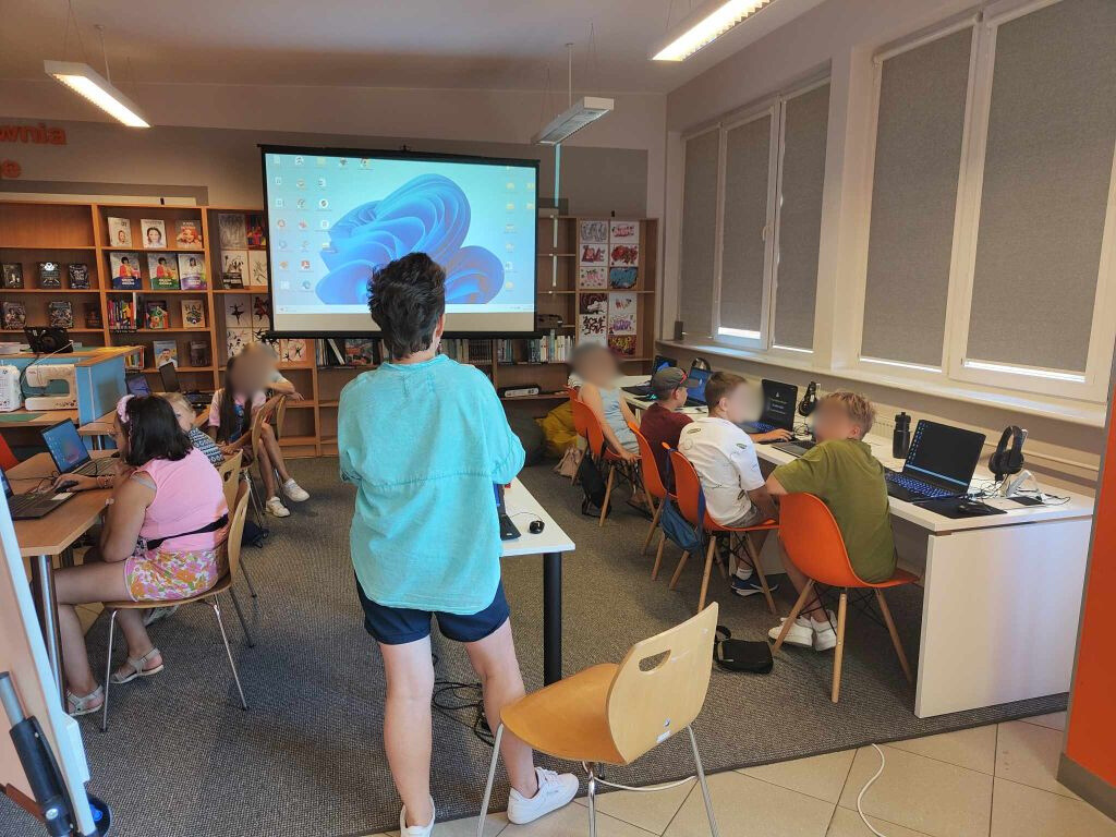 grupa dzieci siedzi przy laptopach, po środku stoi kobieta, w tle ekran projekcyjny z wyświetlanym obrazem