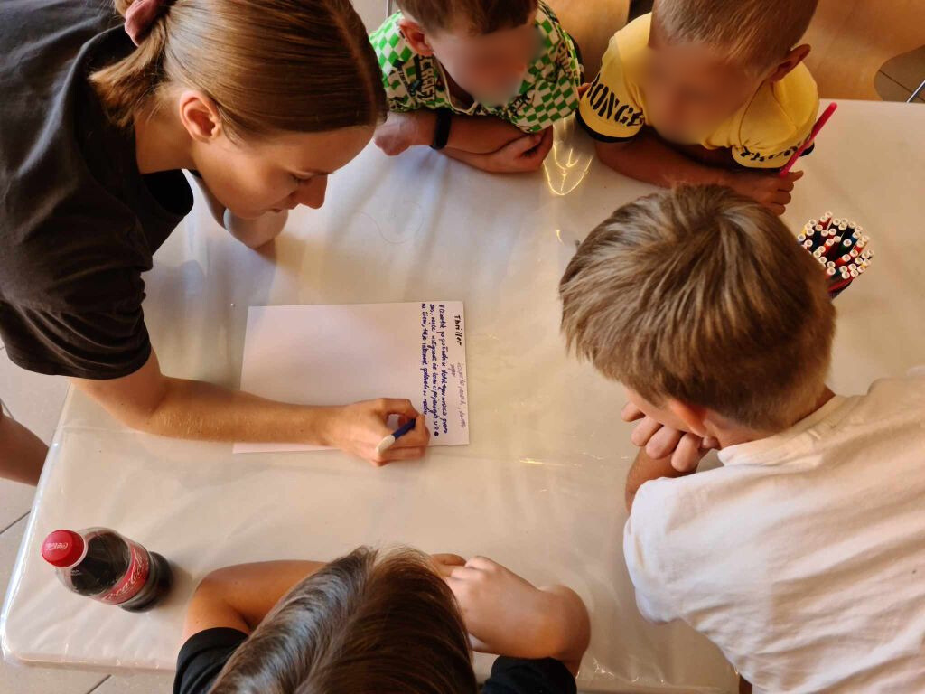 dzieci i kobieta siedzący przy stole i pochylający się nad kartką papieru na której kobieta coś pisze