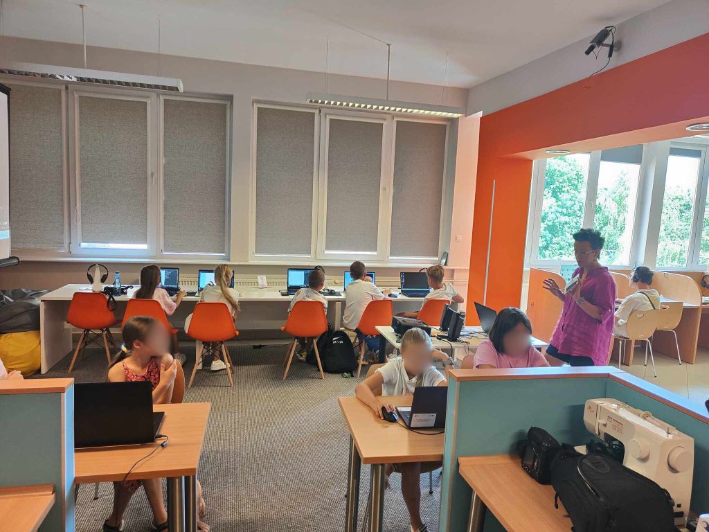 grupa dzieci siedzi przy biurkach przed laptopami, po lewej stoi kobieta