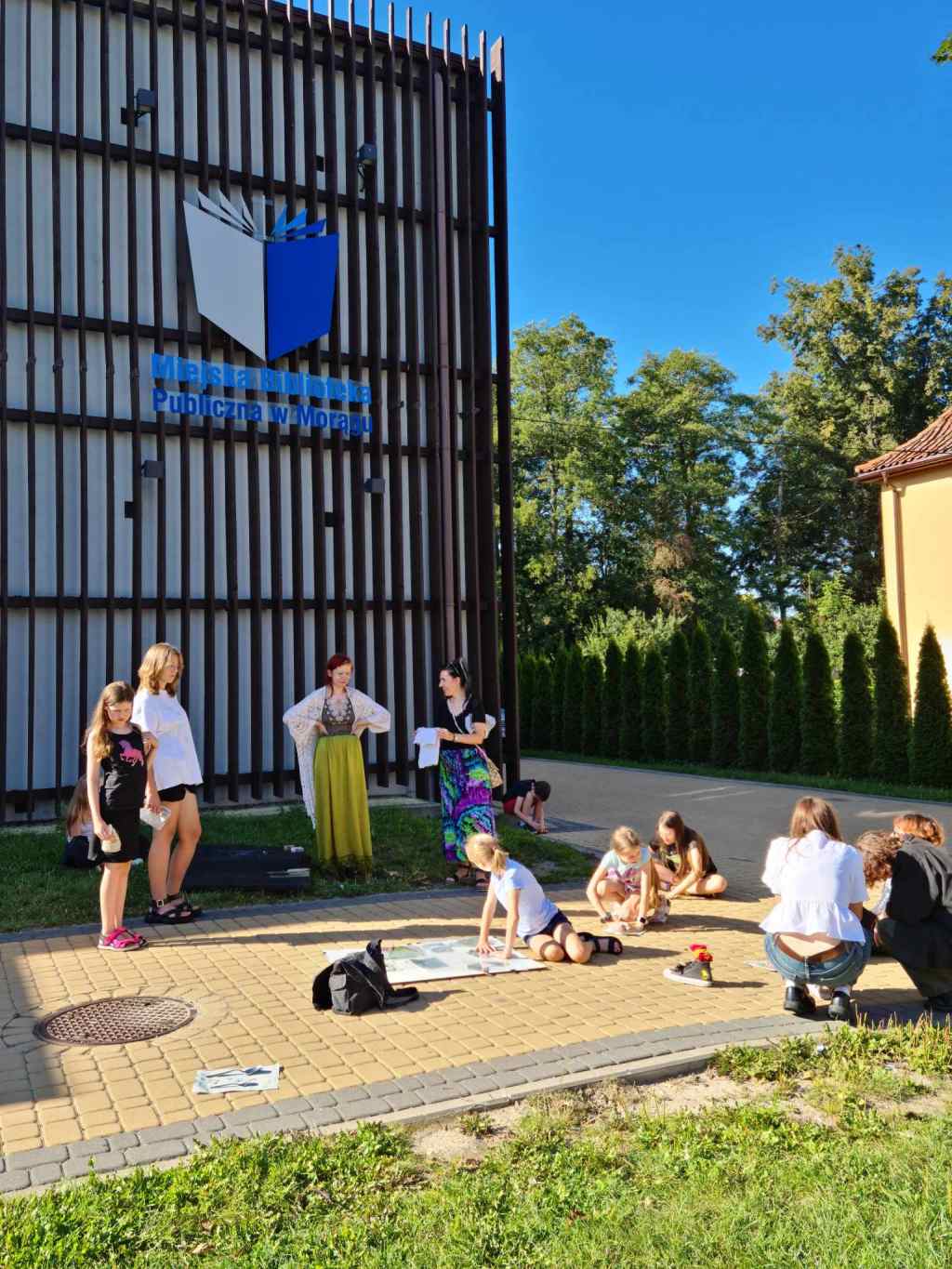 kilka osób siedzi, klęczy lub stoi przed budynkiem biblioteki, na elewacji niebieskie logo z książką