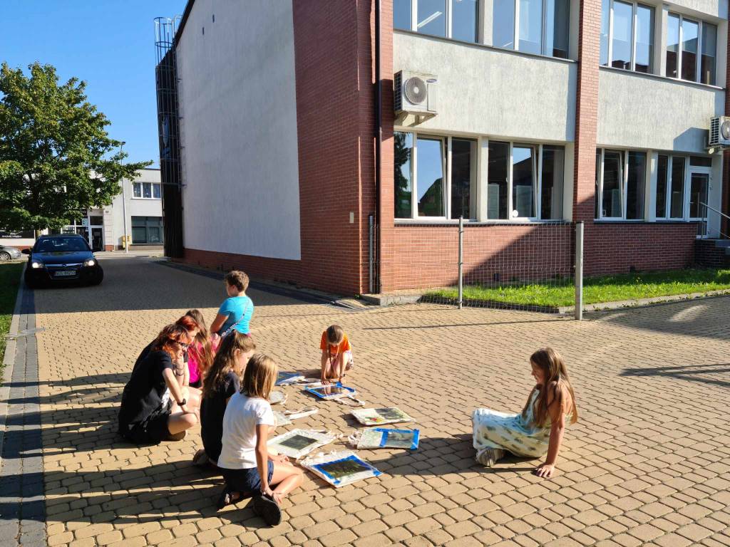 grupa osób siedzi na kostce brukowej, miedzy nimi na ziemi leżą prace plastyczne, za nimi budynek