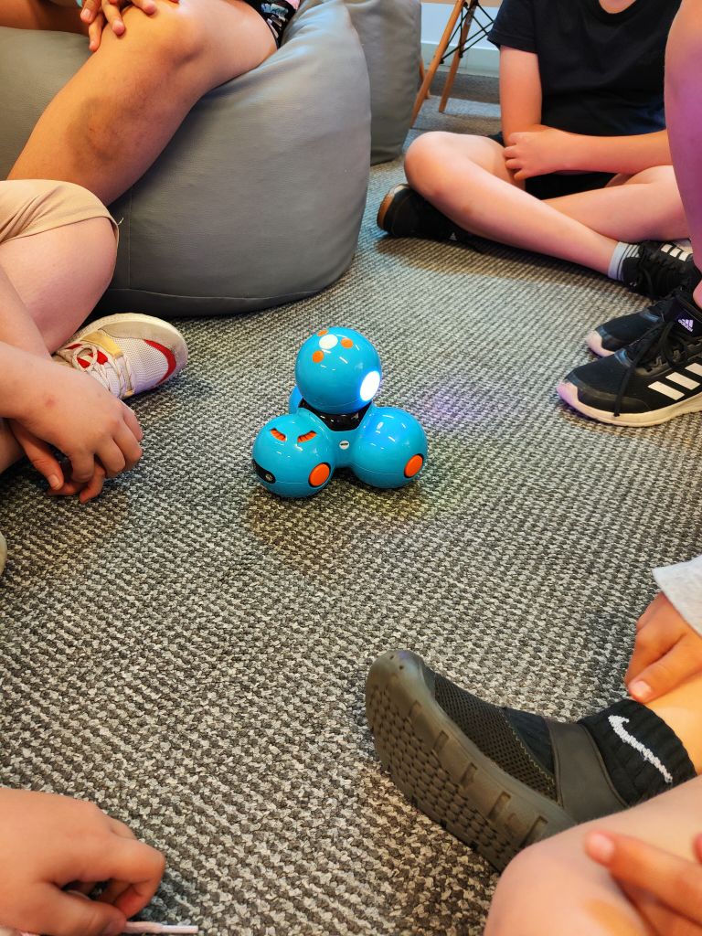 Niebieski robot Dash stoi na podłodze, wokół niego siedzą dzieci