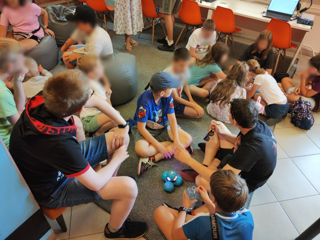 Grupa dzieci siedzi na podłodze, między nimi stoi niebieski robot Dash