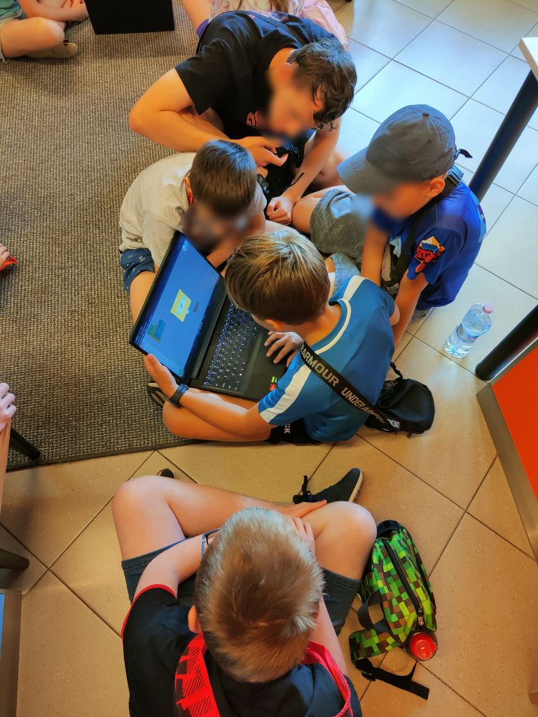 Grupa dzieci siedzi na podłodze, jeden z chłopców trzyma na kolanach laptopa