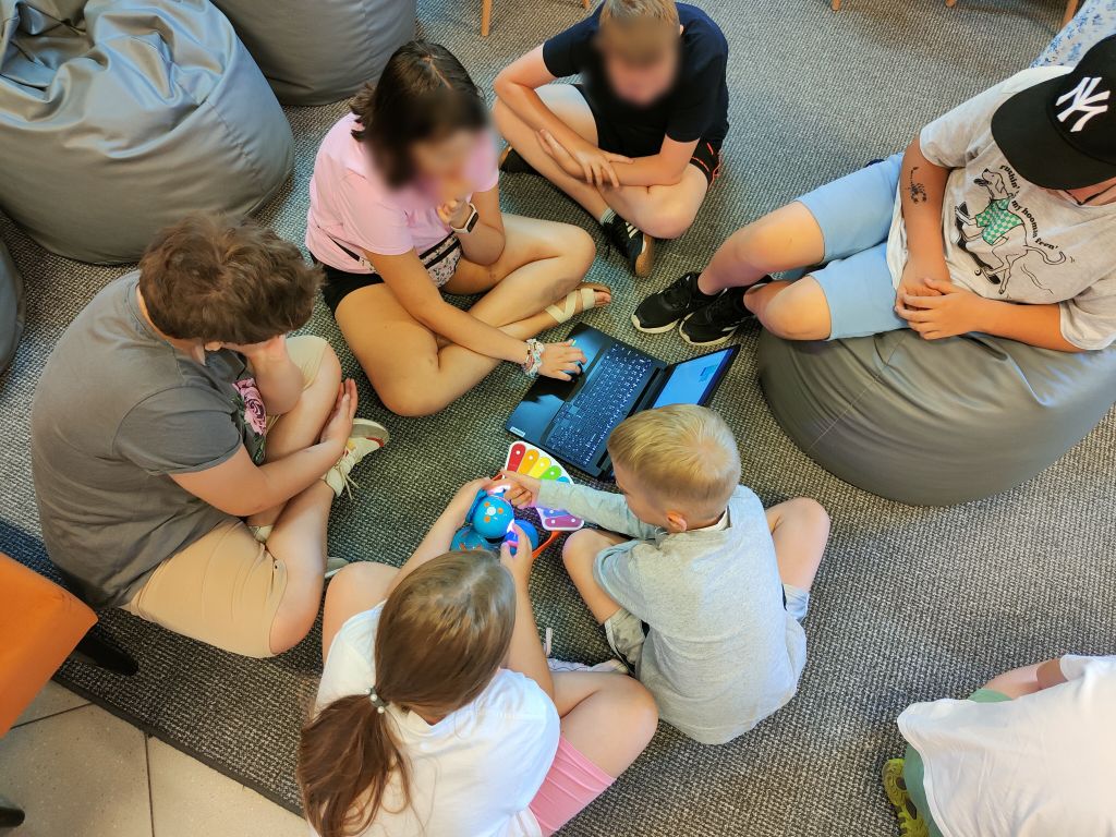 Grupa dzieci siedzi na podłodze, między nimi stoi niebieski robot Dash i laptop