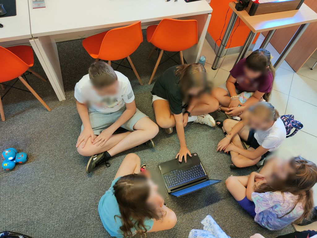 Grupa dzieci siedzi na podłodze, między nimi stoi niebieski robot Dash i laptop