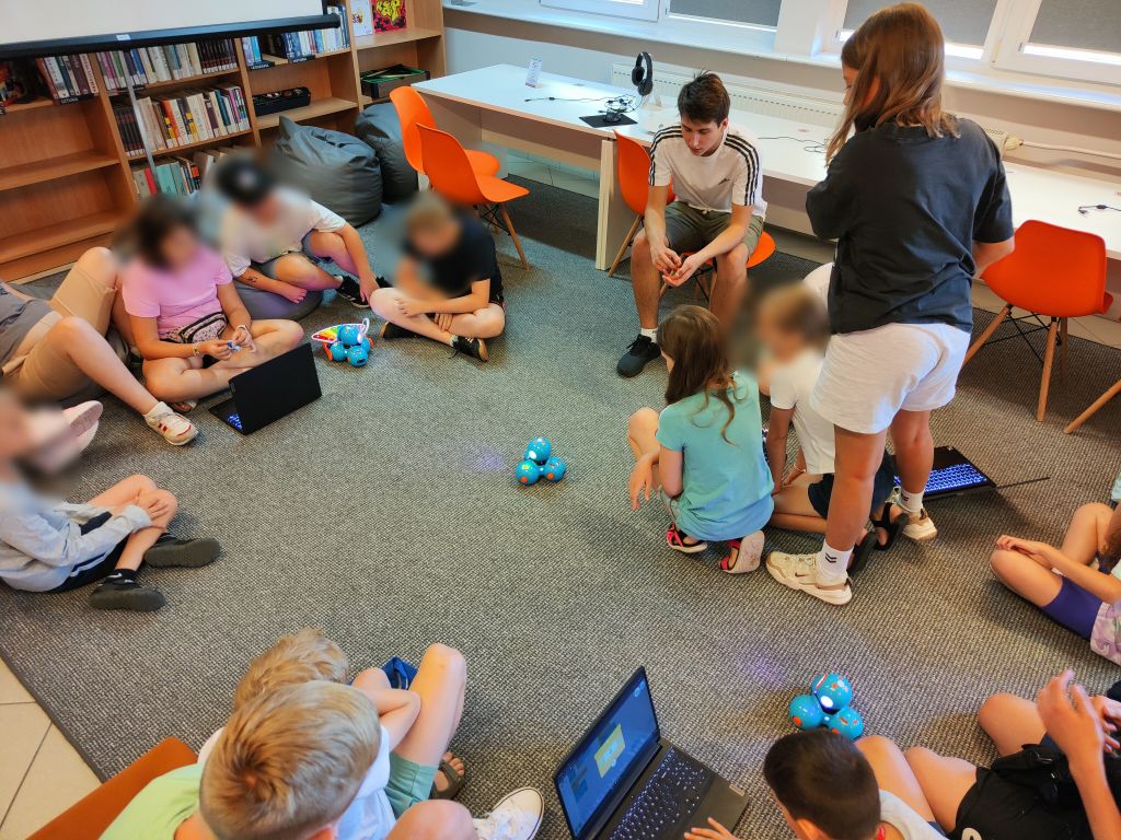 Grupa dzieci siedzi na podłodze, między nimi stoją niebieskie roboty Dash i laptopy