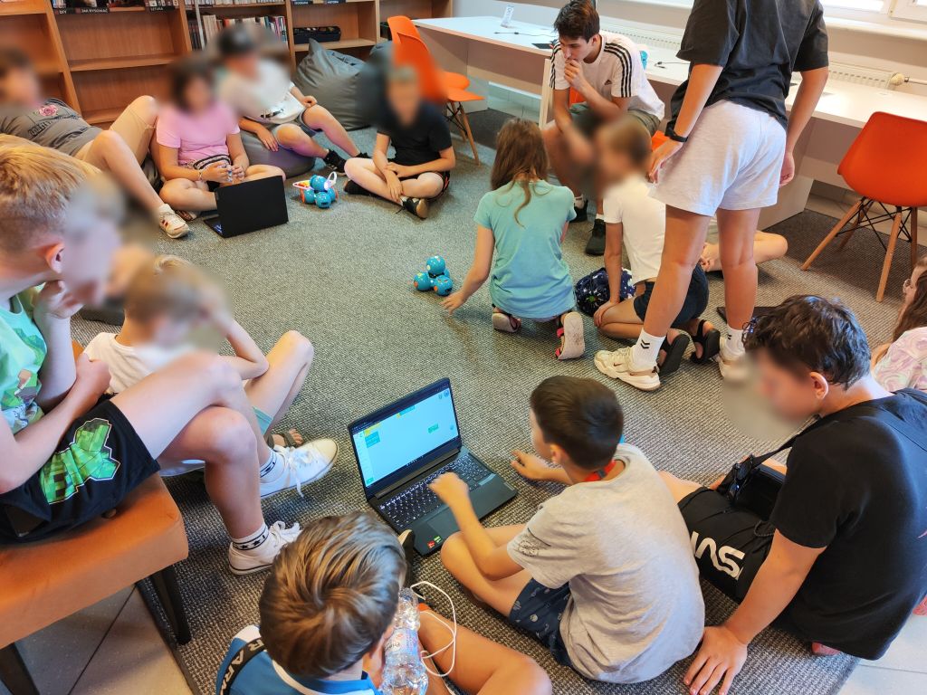 Grupa dzieci siedzi na podłodze, między nimi stoją niebieskie roboty Dash i laptopy