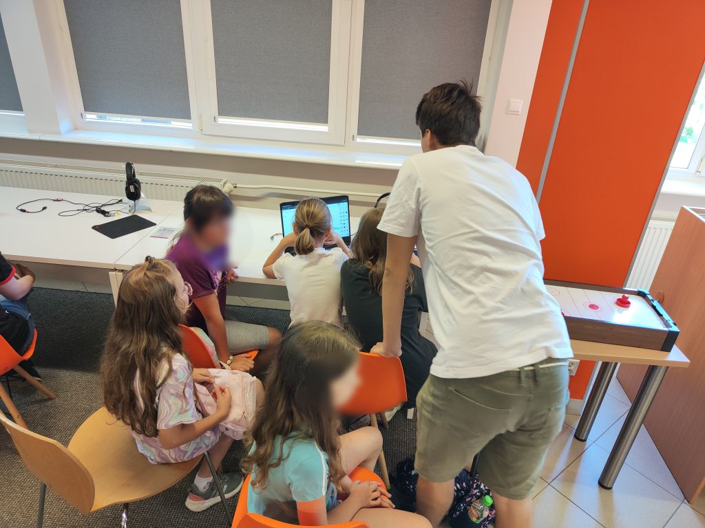 Grupa dzieci siedzi przy biurku, jedno z nich korzysta z laptopa. Nad grupą pochyla się mężczyzna