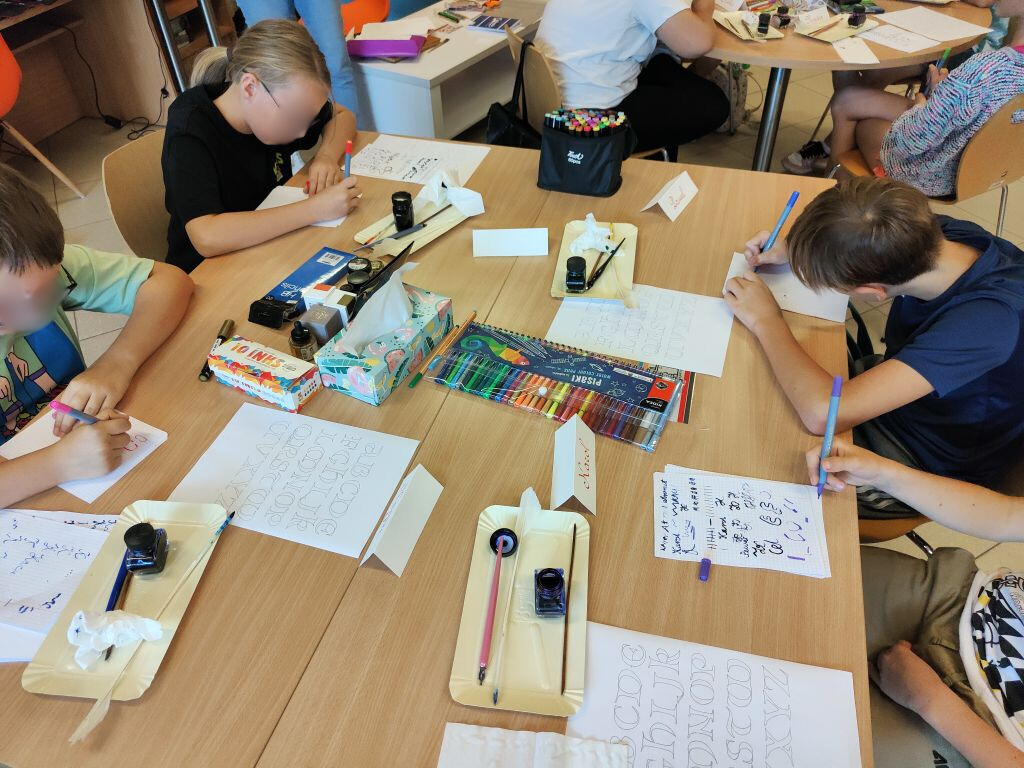 Dzieci siedzą przy stołach i ćwiczą kaligrafię. Na stołach widoczne materiały plastyczne do kaligrafii