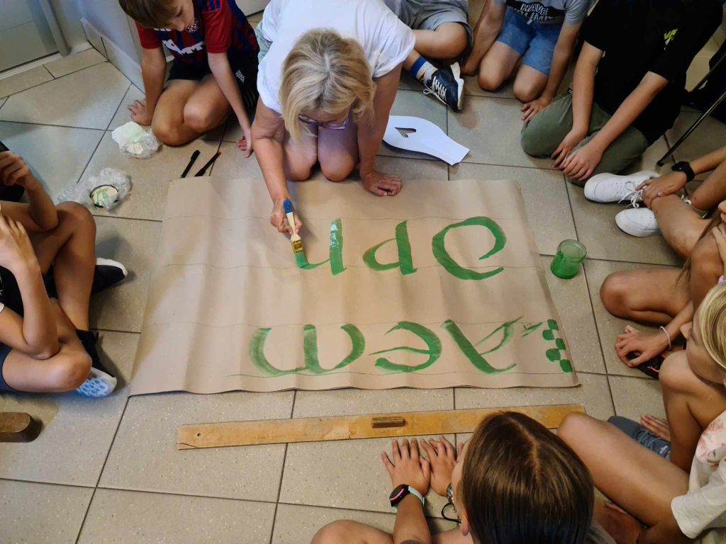 kobiety pochyla się nad dużym arkuszem papieru rozłożonym na podłodze, na którym maluje litery. Wokół niej siedzi grupa dzieci