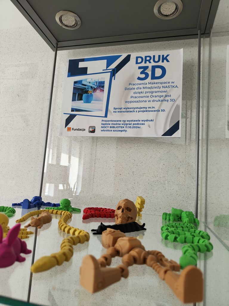 Gadżety wydrukowane na drukarce 3D ułożone na półce w gablocie. W tle plakat promujący wystawę powieszony na tylnej ścianie gabloty