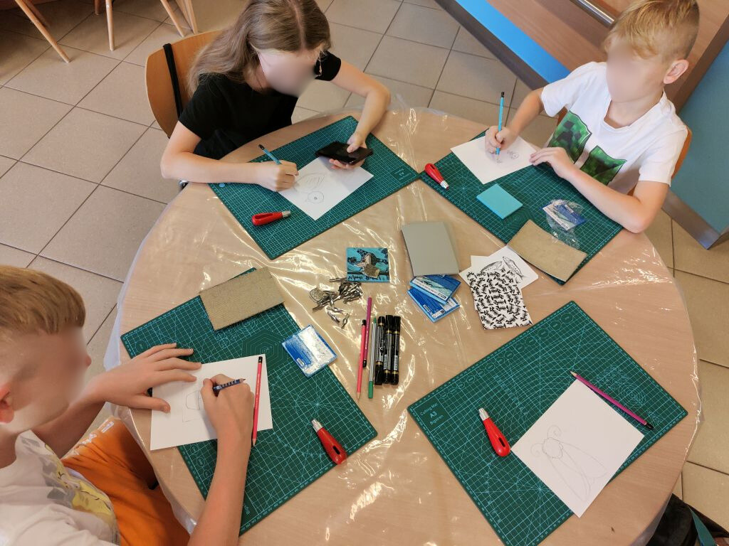 cztery osoby siedzą przy okrągłym stole i przygotowują negatywy linorytów w linoleum