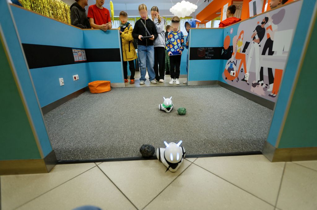 na podłodze 2 małe, białe roboty, za nimi dwoje dzieci trzyma tablety i steruje robotami, za nimi grupka kibiców