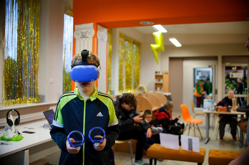 chłopiec ma założone na głowie gogle VR, w rękach trzyma pady, za nim w tle siedzi kilka osób