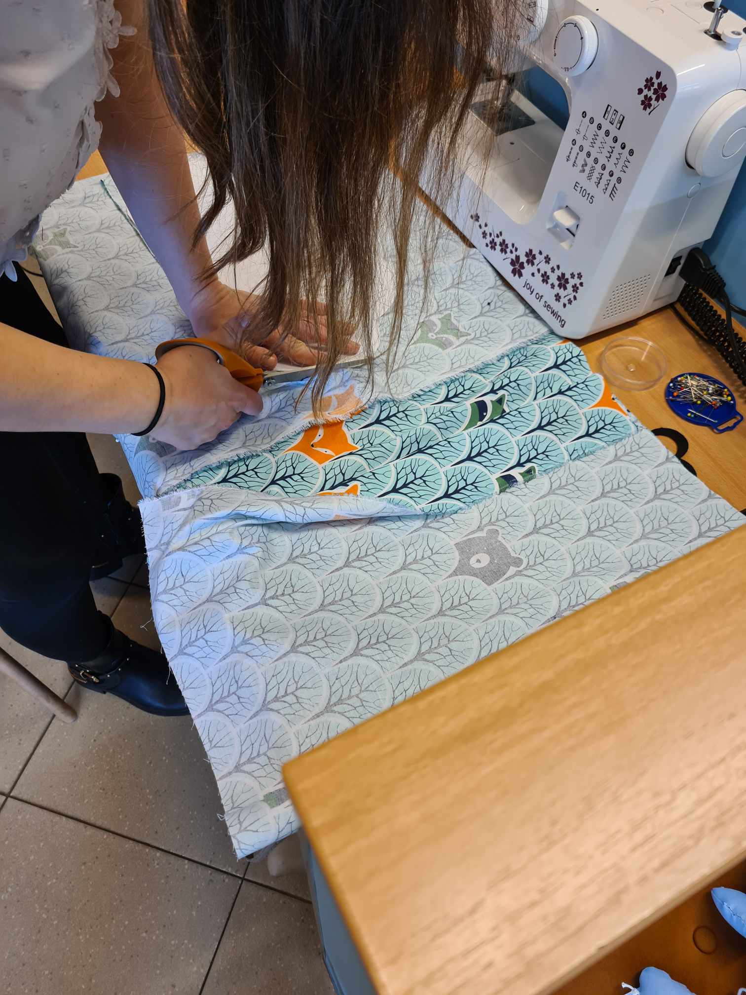 kobieta nachyla się nad stołem, kroi materiał, przed nią stoi maszyna do szycia
