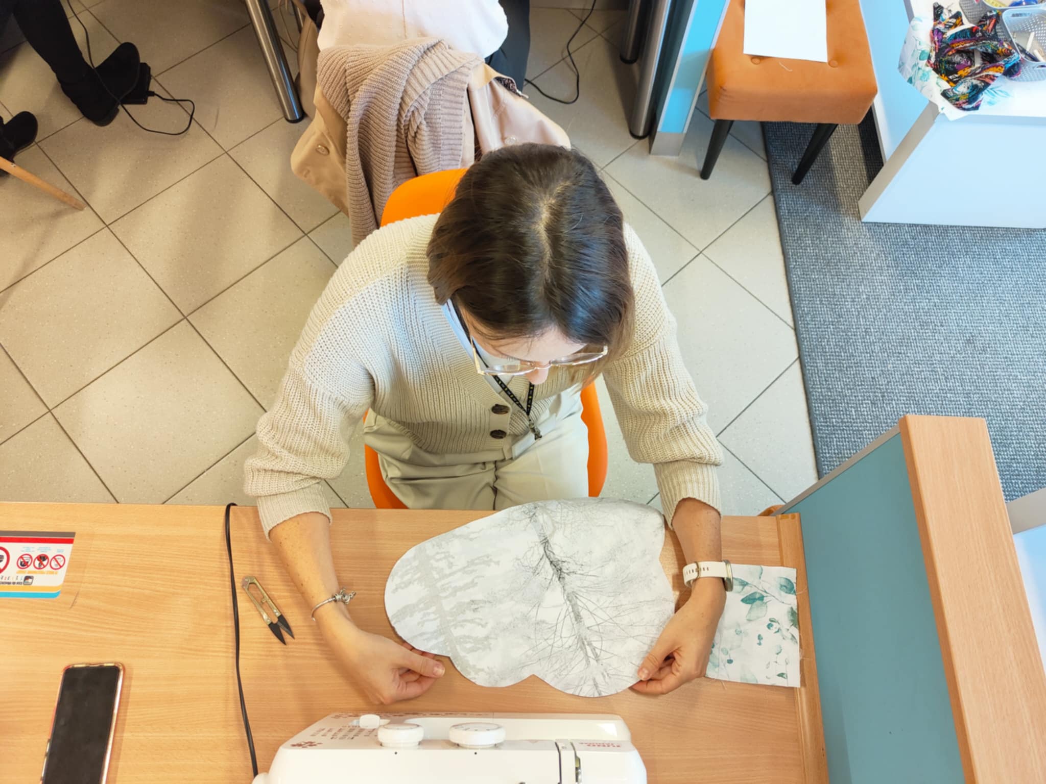 kobieta siedzi przy stole przed maszyną do szycia, w rękach trzyma chmurę wycięta z tkaniny