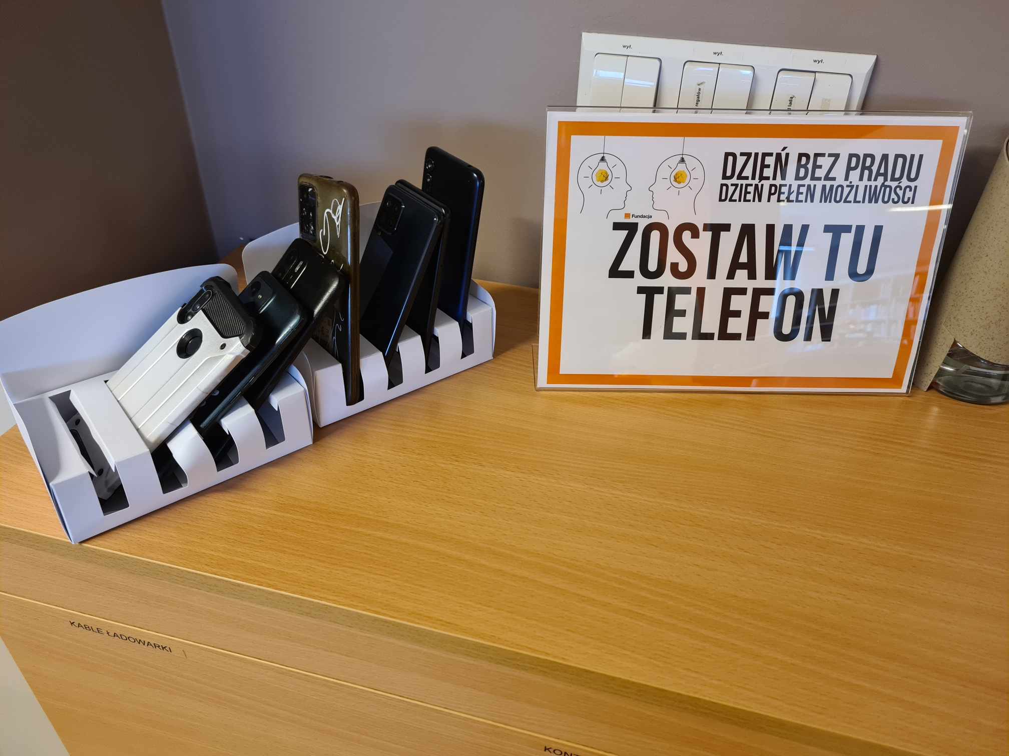 na szafce kartka z napisem Zostaw tu telefon, obok stoi kilka telefonów