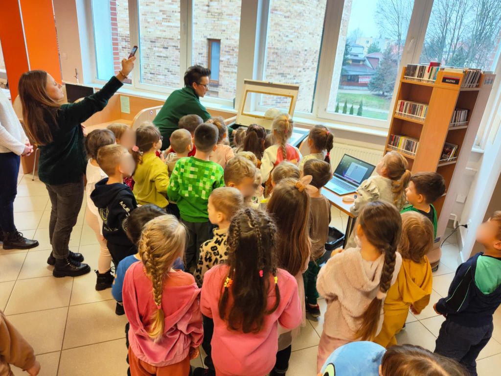 dzieci stoją na przeciwko plotera laserowego i patrzą na to co pokazuje im stojąca obok plotera kobieta