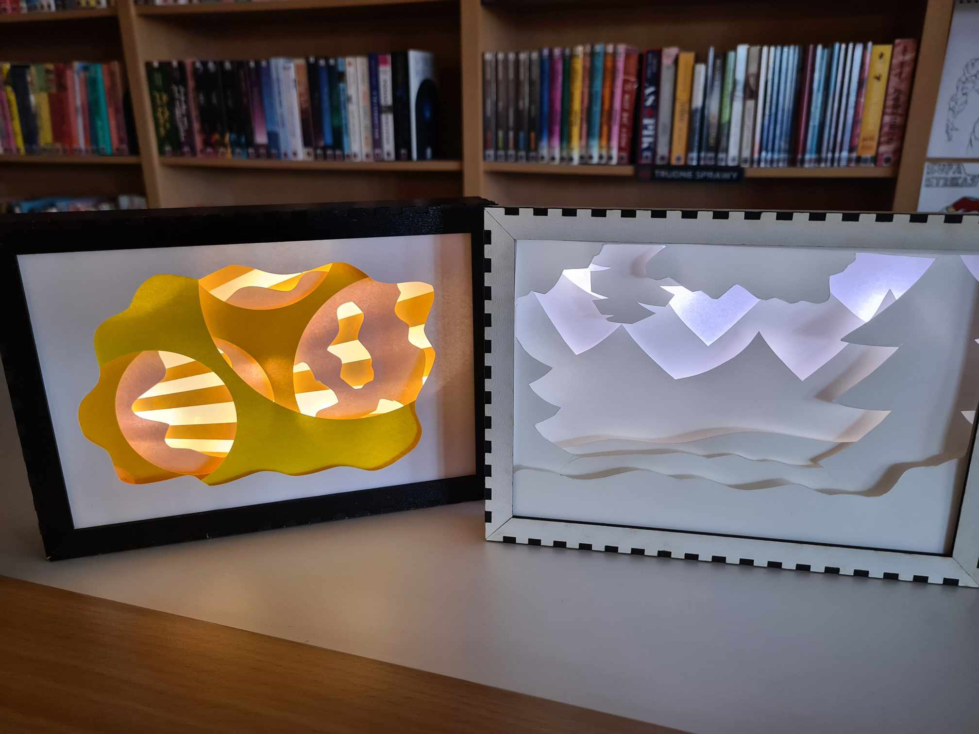 Dwa pudełka cieni, jedno kolorowe z kształtami geometrycznymi, drugie białe z pejzażem, w tle regały z książkami