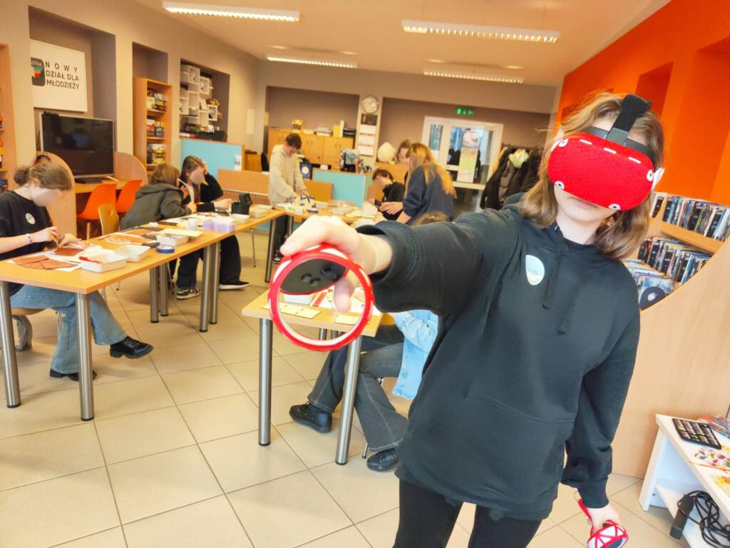 na pierwszym planie dziewczyna korzystająca z gogli VR, w tle grupa nastolatków biorąca udział w warsztatach