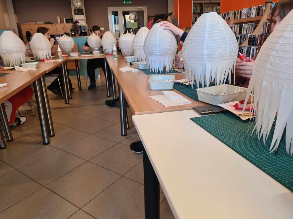 grupa osób siedzi przy stołach i przyklejają białe laski papiery do kloszów lampek