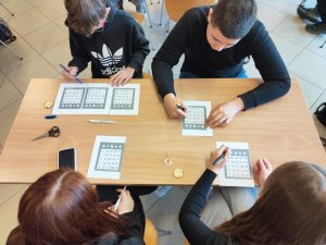 Na zdjęciu widać cztery osoby siedzące przy prostokątnym stole, które grają w bingo. Na stole znajdują się również nożyczki, smartfon oraz mały okrągły przedmiot, być może żeton lub moneta.