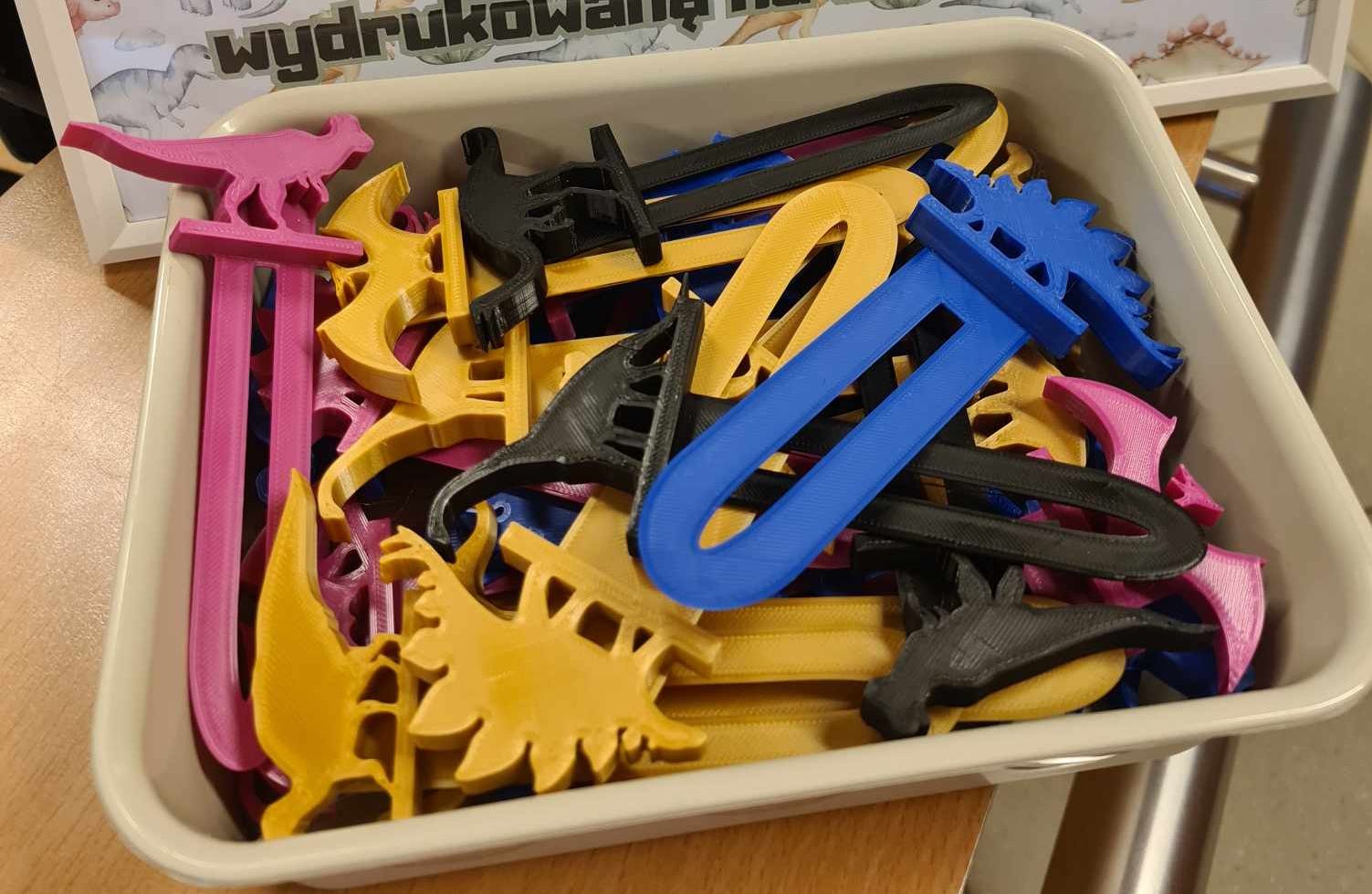 Na zdjęciu znajduje się plastikowy pojemnik wypełniony kolorowymi zakładkami do książek w kształcie dinozaurów. Zakładki są wykonane z tworzywa sztucznego i mają różne kolory: różowy, żółty, czarny i niebieski. Każda zakładka ma na górze figurkę dinozaura. W tle widoczny jest fragment plakatu z napisem "wydrukowaną", co sugeruje, że zakładki mogły zostać wydrukowane za pomocą drukarki 3D.