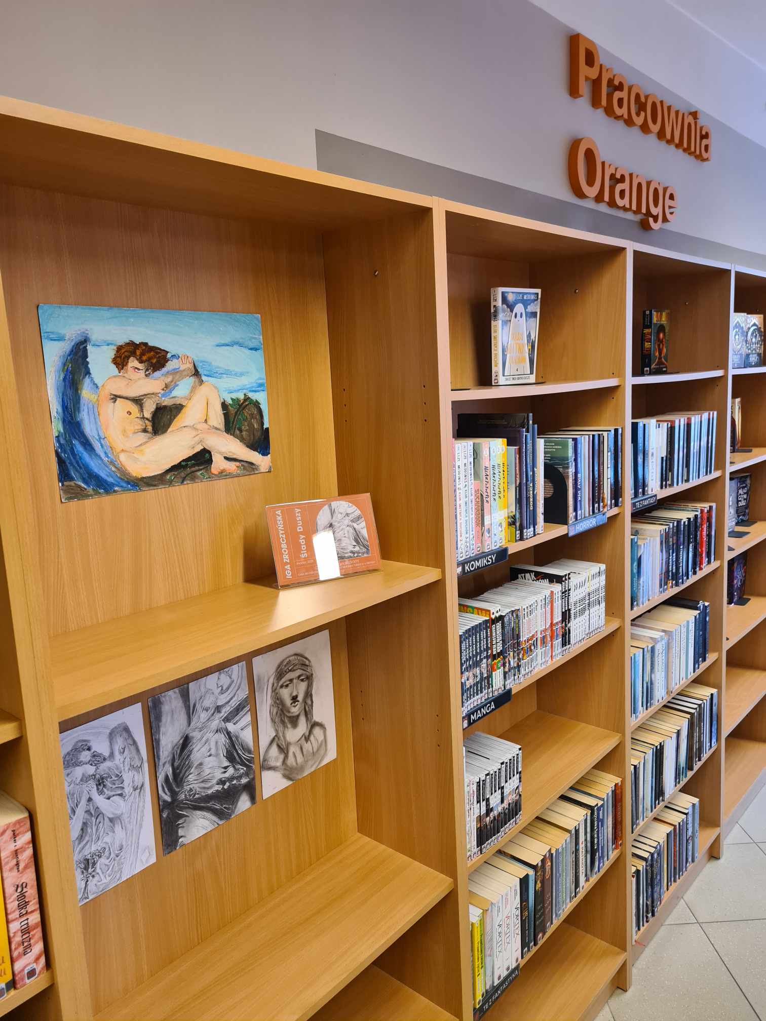 Biblioteka z drewnianymi regałami wypełnionymi różnymi książkami. Na górze obrazu widnieje napis „Pracownia Orange”. Półki zawierają mieszankę książek, w tym mangi i inne gatunki. Na regałach umieszczone są również dzieła sztuki, takie jak obraz osoby siedzącej na tle niebieskim oraz trzy czarno-białe szkice.