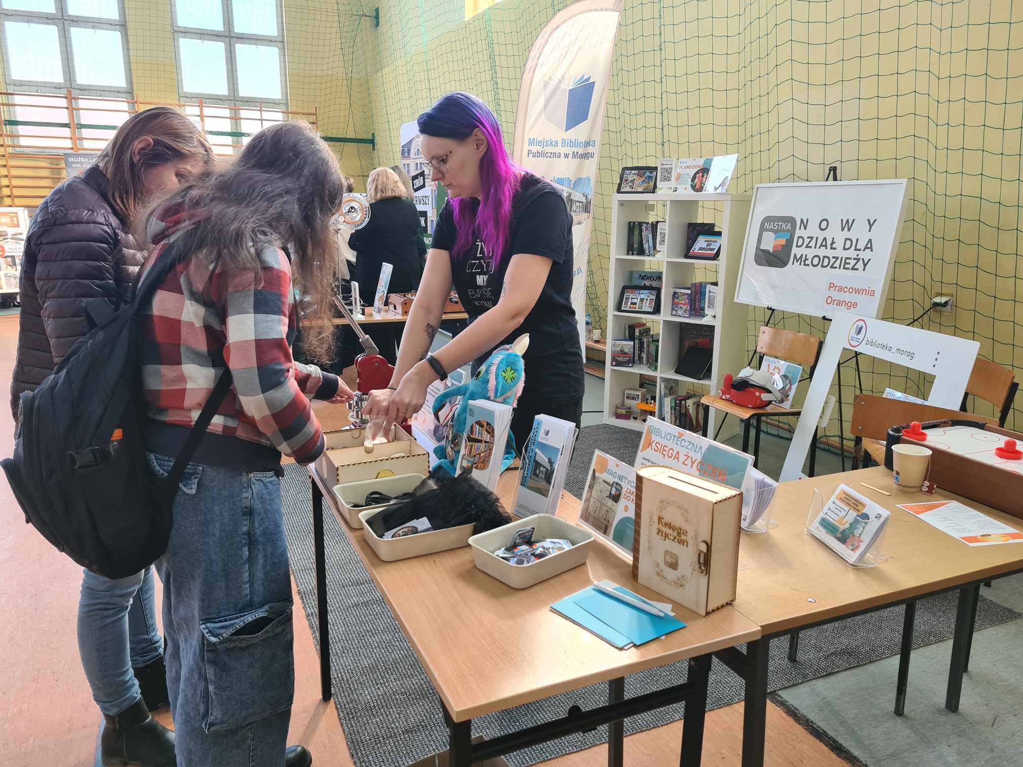 Na zdjęciu widać stoisko na targach, które odbywa się w sali gimnastycznej. Na pierwszym planie znajduje się stół z różnymi przedmiotami, takimi jak książki, broszury, gadżety i materiały promocyjne. Po prawej stronie stołu stoi osoba z fioletowymi włosami, ubrana w czarną koszulkę, która pomaga dwóm innym osobom przyglądającym się przedmiotom na stole. W tle widać regał z książkami oraz banery promocyjne. Jeden z banerów zawiera napis 