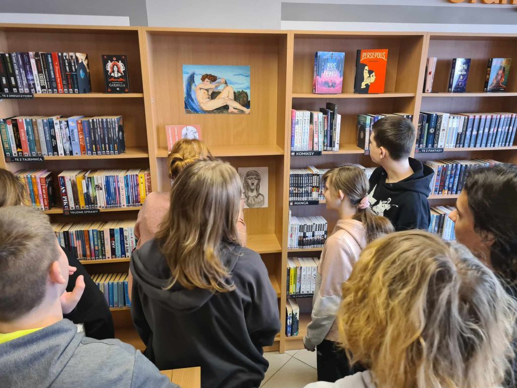 Grupa osób stoi przed regałem z książkami w bibliotece. Regał jest wypełniony różnorodnymi książkami, w tym z gatunków takich jak fantasy, manga i komiksy. Na półkach znajdują się także rysunki, w tym obraz osoby leżącej oraz czarno-biały portret. Osoby oglądają regał, prawdopodobnie dyskutując na temat sztuki.