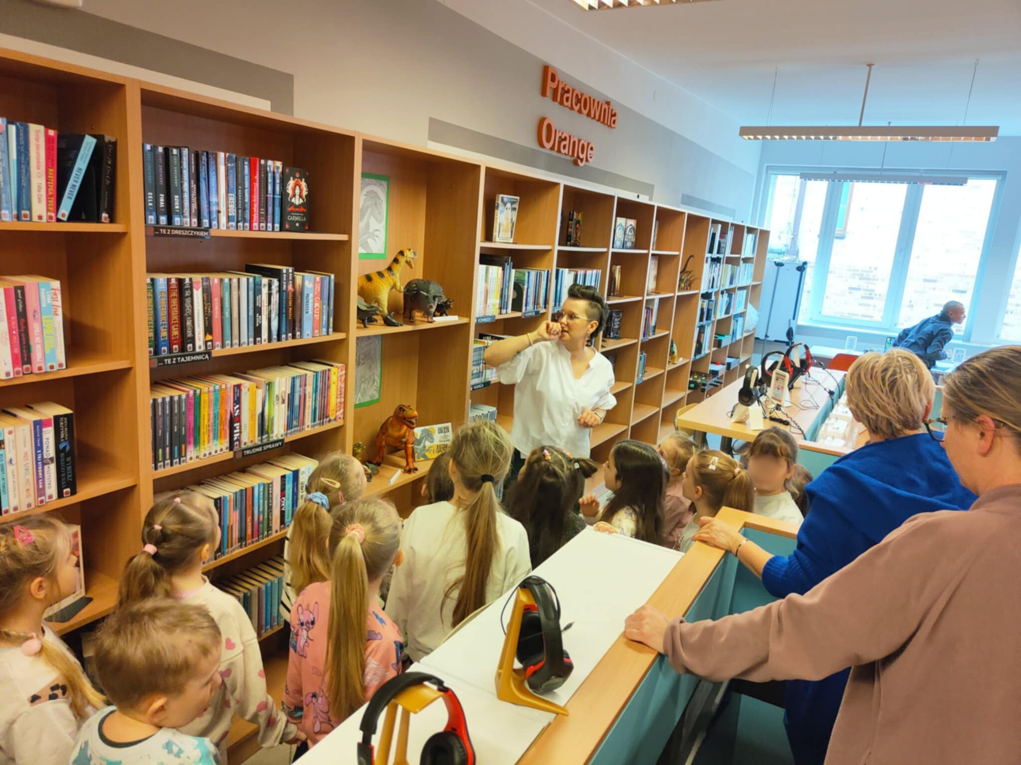 Na zdjęciu widać grupę dzieci i dorosłych w bibliotece, stojących przed półkami z książkami i zabawkami w kształcie dinozaurów. Pomieszczenie jest jasne, dzięki dużym oknom wpuszczającym światło dzienne. W tle widać regały z książkami.