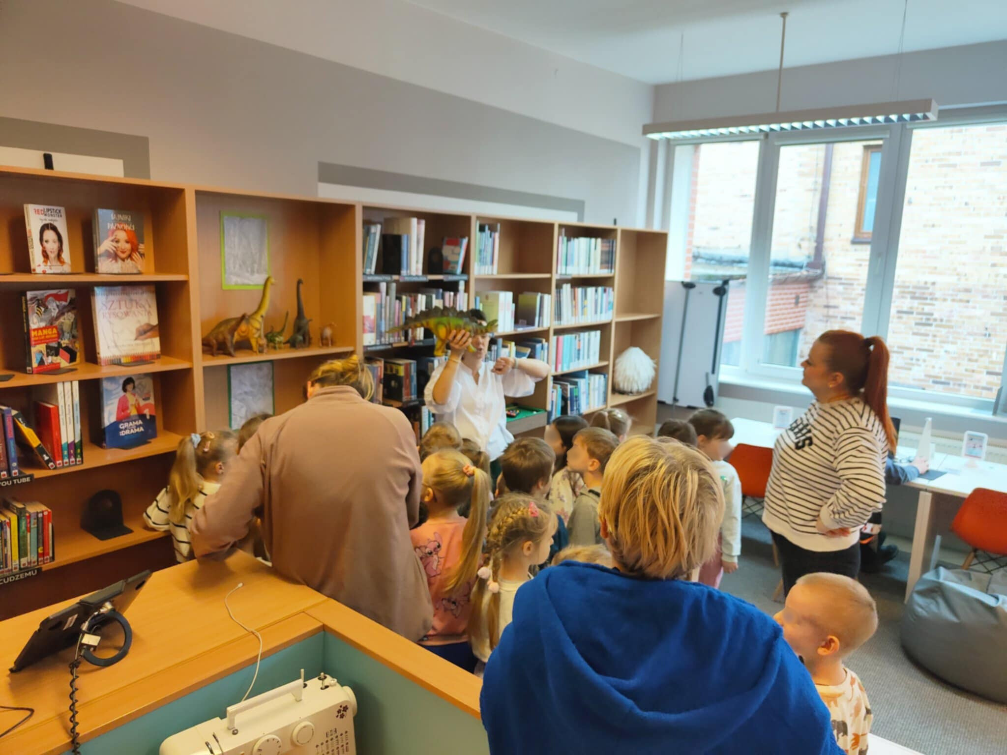 Na zdjęciu widać grupę dzieci i dorosłych w bibliotece, stojących przed półkami z książkami i zabawkami w kształcie dinozaurów. Pomieszczenie jest jasne, dzięki dużym oknom wpuszczającym światło dzienne. W tle widać regały z książkami. Kobieta Trzyma w ręku dinozaura