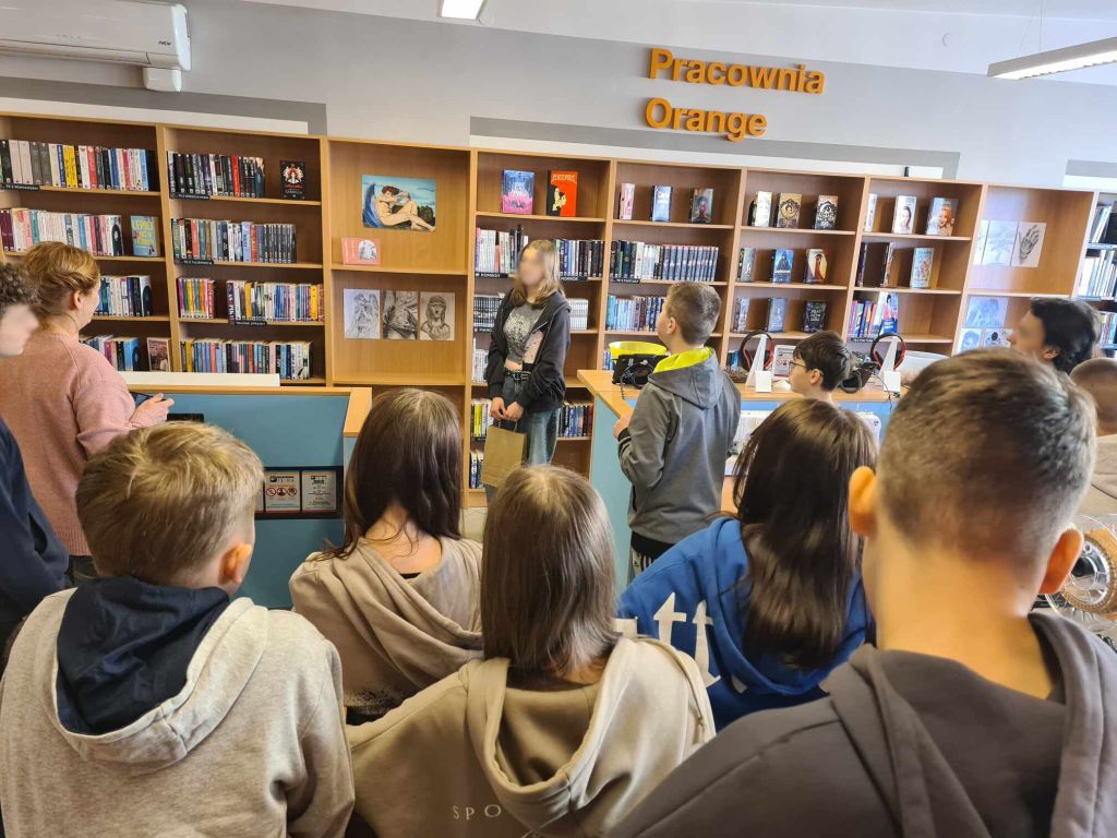 Grupa uczniów, zgromadzona w bibliotece. W tle widać regały wypełnione książkami, nad nimi na ścianie napis Pracownia Orange, a na niektórych półkach wyeksponowane są dzieła sztuki. Osoby stoją i wydają się słuchać kogoś, kto mówi. Scena sugeruje zwiedzanie galerii.