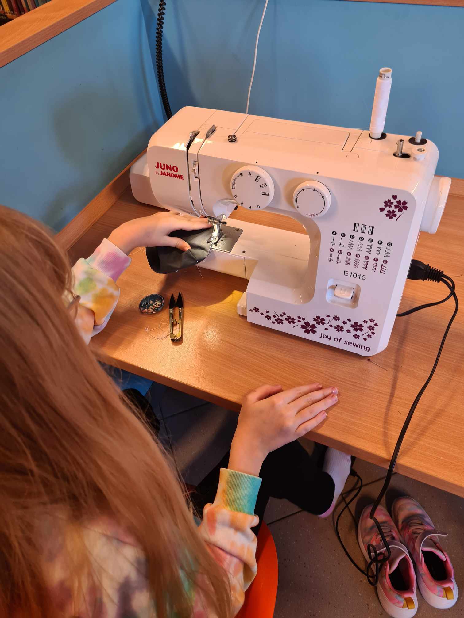 Dziewczynka z długimi blond włosami szyje na białej maszynie do szycia marki Janome. Trzyma czarny materiał, prowadząc go pod igłą. Na biurku obok maszyny leżą nożyczki, szpulka nici i akcesoria krawieckie. Przy ścianie stoi para różowych butów sportowych.