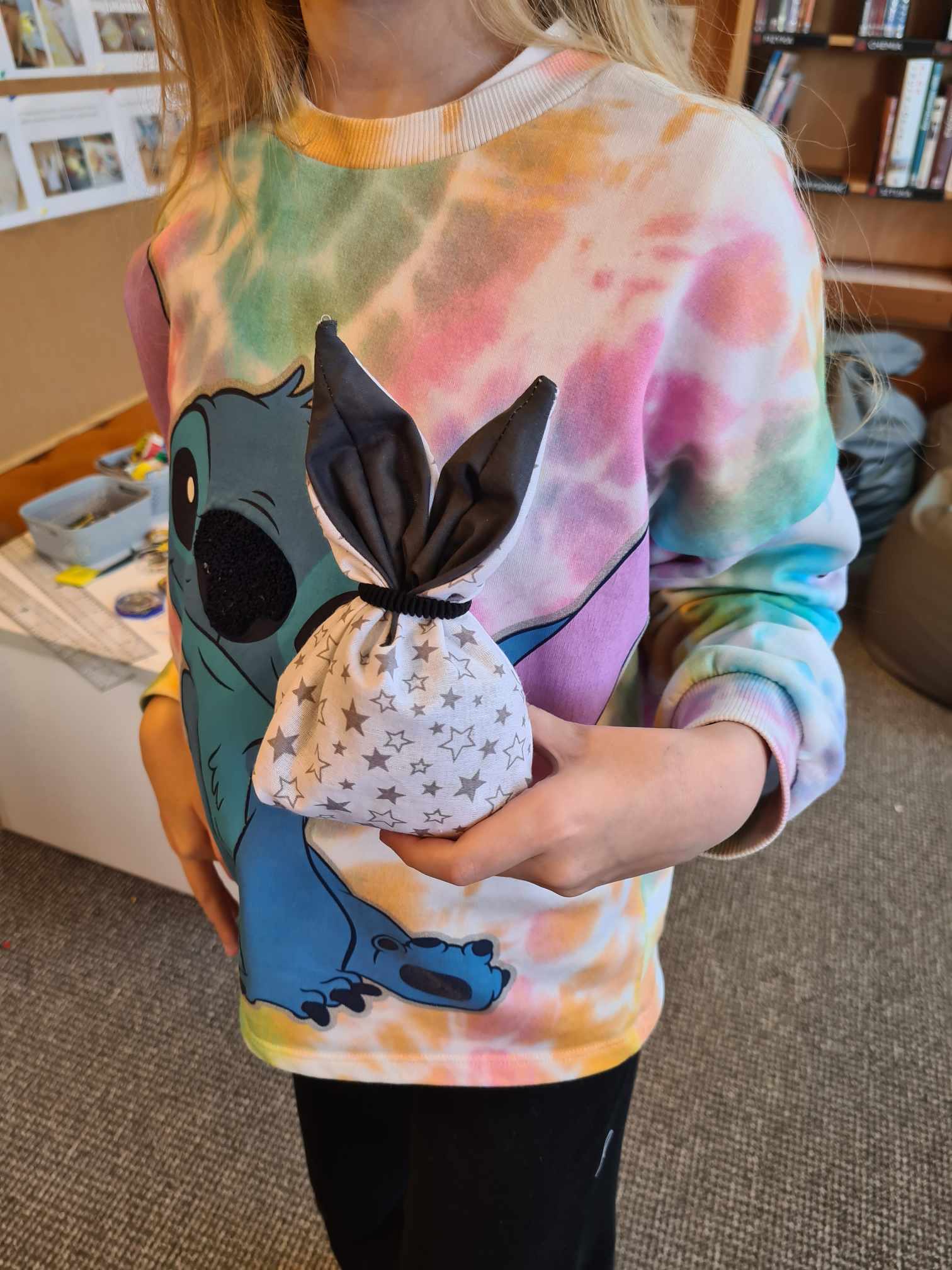 Dziewczynka w kolorowej bluzie z postacią Stitcha trzyma w dłoniach materiałowy koszyczek w kształcie króliczka. Koszyczek jest biały w szare gwiazdki, a jego uszy są czarne z białym obszyciem. W tle widać biblioteczne regały i stolik z akcesoriami do szycia.