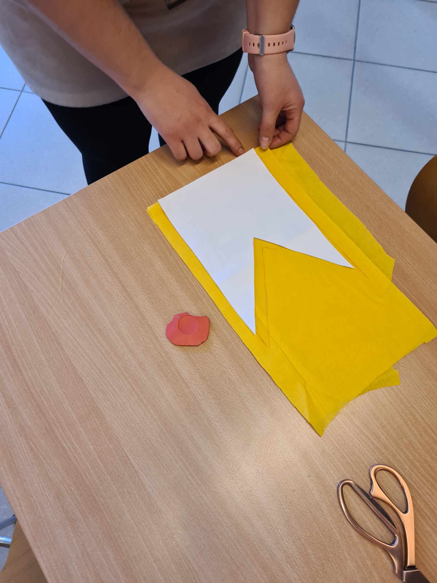 Osoba pracuje nad projektem rękodzielniczym. Na żółtym materiale układa biały papier z wyciętym trójkątem.
