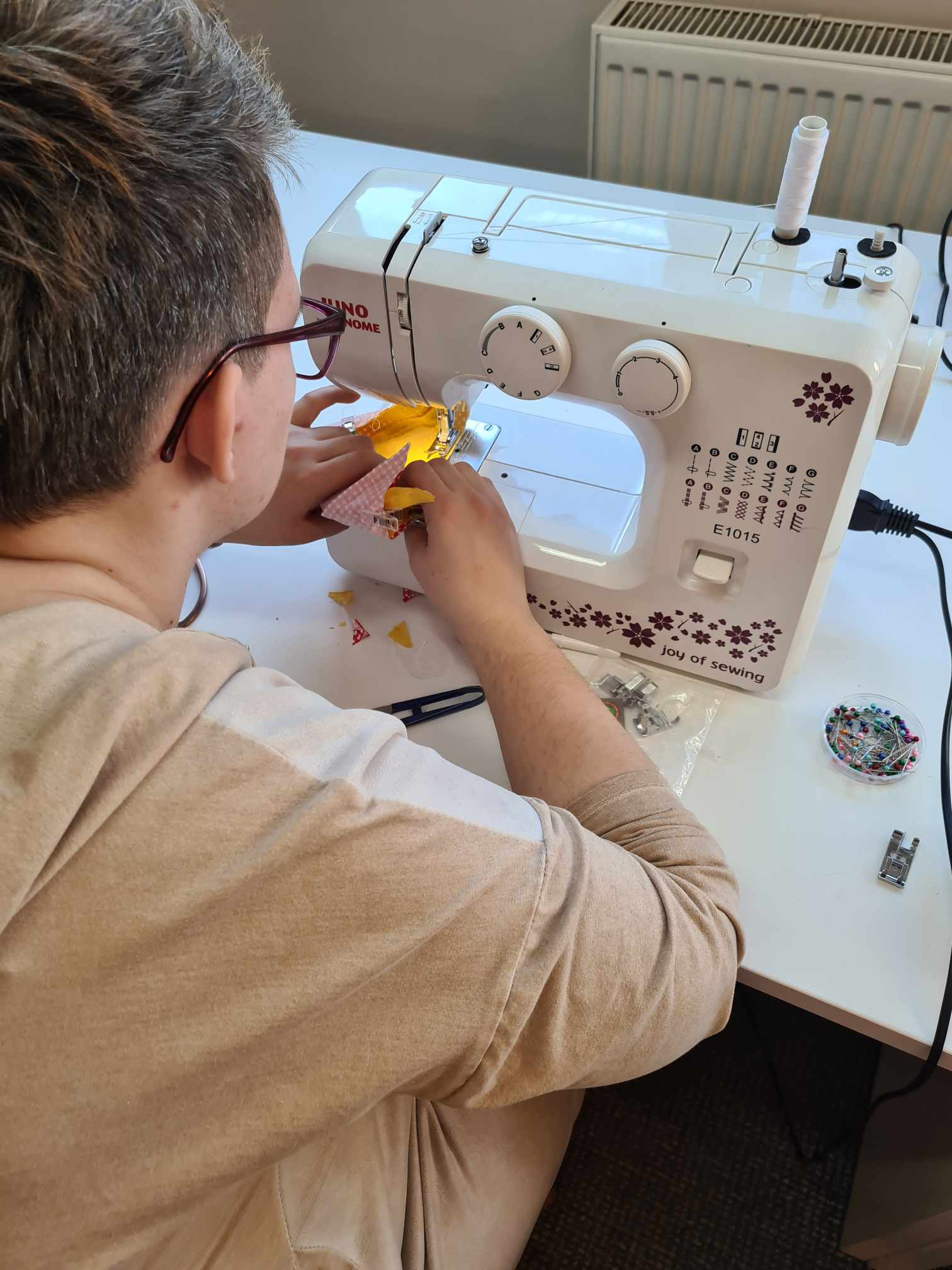 Zdjęcie przedstawia osobę korzystającą z maszyny do szycia, modelu Janome E1015, która jest biała z kwiatowymi ozdobami i napisem „joy of sewing”. Osoba szyje kawałek materiału, który wydaje się być żółto-różowy. Na stole znajdują się akcesoria do szycia, takie jak pudełko ze szpilkami, nożyczki i stopki do maszyny.