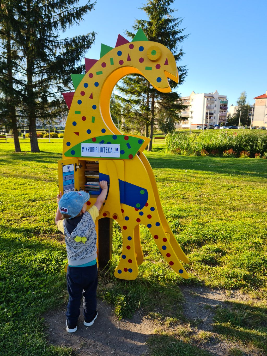 W parku znajduje się kolorowa konstrukcja w kształcie dinozaura, oznaczona napisem „MIKROBIBLIOTEKA.” Dinozaur pełni funkcję punktu wymiany książek. Jej korpus jest żółty i ozdobiony różnokolorowymi kształtami – kropkami i trójkątami. Przed biblioteką stoi dziecko, które sięga do wnętrza konstrukcji. W tle widoczne są zielona trawa, drzewa oraz budynki.